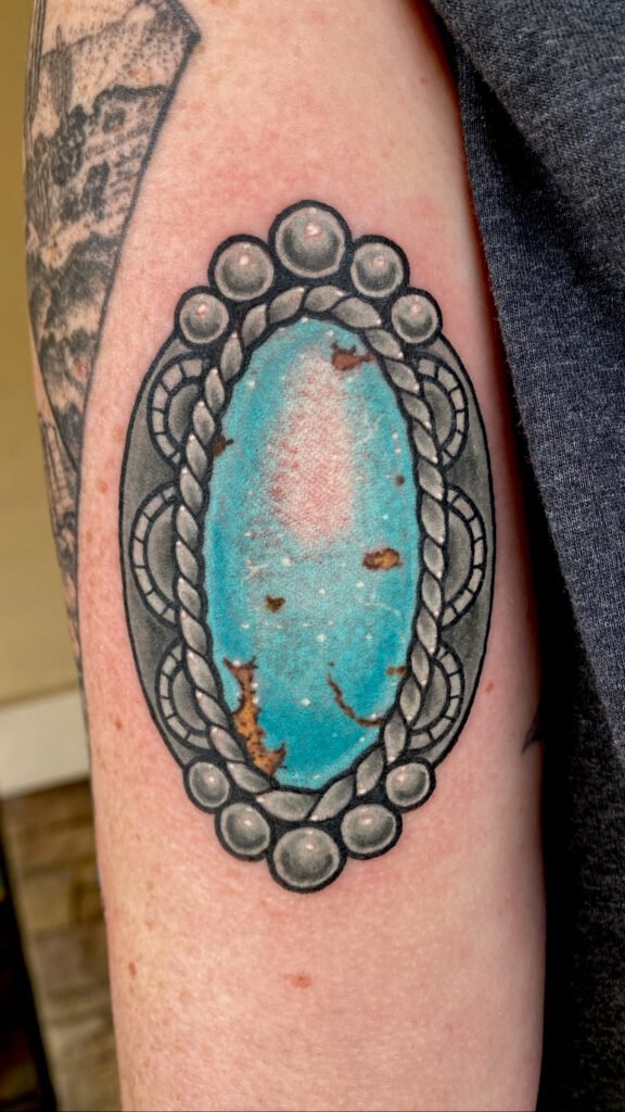 a neo traditional tattoo of a piece of turquoise and silver jewelry. The turquoise jewel gem has brown specs to resemble natural impurities in the mineral. It has an ornate frame resembling aged silver ink. This tattoo was done by Jeremy Moffitt an artist at Near Me Tattoo located at 6721 Bells Ferry Rd Ste D110 Woodstock, Ga. 30189