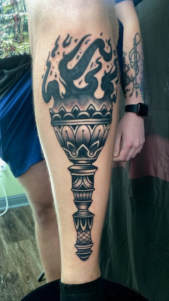 A Neo traditional torch tattoo on the entire shin. It is done in blackwork style shading. This gives a reverse fade to the flames coming from the goblet of the torch. The handle is ornate and bold. This tattoo was done by Jeremy Moffitt an artist at Near Me Tattoo located at 6721 Bells Ferry Rd Ste D110 Woodstock, Ga. 30189