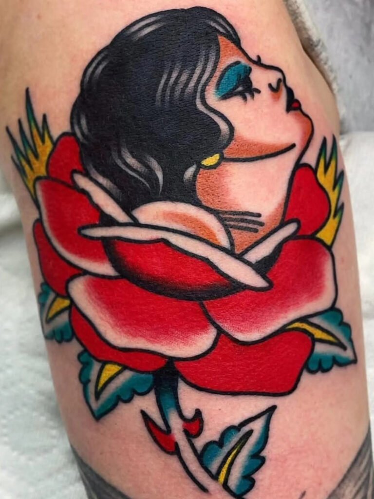 An American traditional girl head coming out of a rose. The woman has black hair and teal eyeshadow with a golden earring. The rose is a bright red. This tattoo was made by Noah Beach, co owner of Near Me Tattoo at 6721 Bells Ferry Rd Ste D110 Woodstock, Ga. 30189
