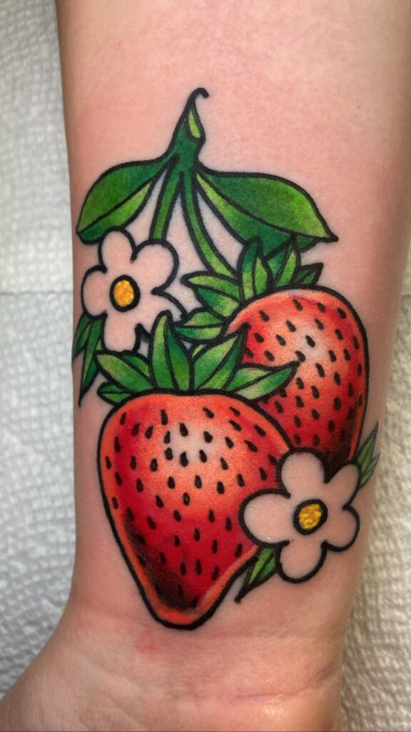A simple tattoo on the wrist of two strawberries and their white flowers. They are attached together at the vine. It has a simple ink palette of black green red and yellow. This tattoo was done by Jeremy Moffitt an artist at Near Me Tattoo located at 6721 Bells Ferry Rd Ste D110 Woodstock, Ga. 30189