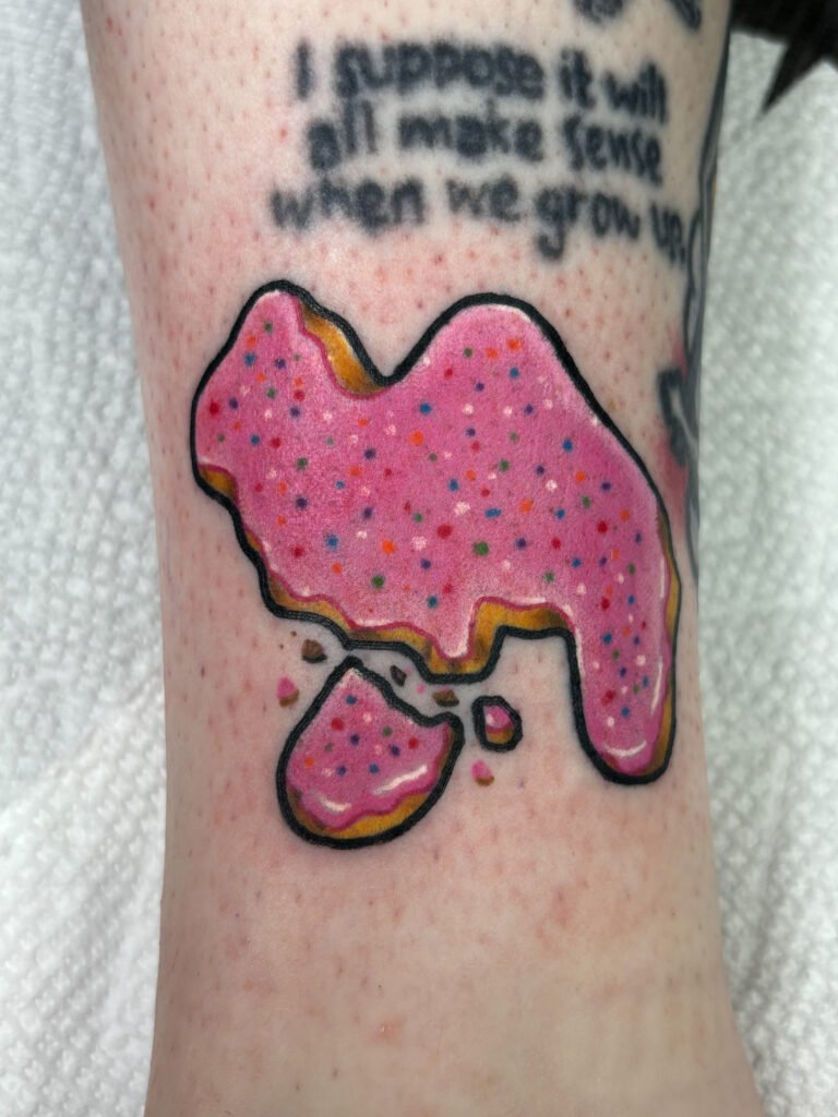 a tattoo of a frosted animal cracker. It has a black bold outline and is brightly colored with pink frosting and multicolored sprinkles. One of the legs has crumbled off. This tattoo was done by Jeremy Moffitt an artist at Near Me Tattoo located at 6721 Bells Ferry Rd Ste D110 Woodstock, Ga. 30189