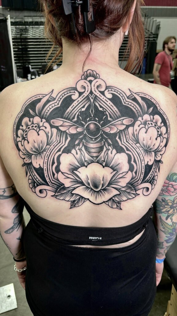 a blackwork tattoo of a full upper back on a woman. It has a bee over a lotus flower and a lotus flower to either side of the bee. It has an ornate semi frame around the sides and top of the tattoo. The heaviest shading is done with solid black ink. This tattoo was done by Jeremy Moffitt an artist at Near Me Tattoo located at 6721 Bells Ferry Rd Ste D110 Woodstock, Ga. 30189