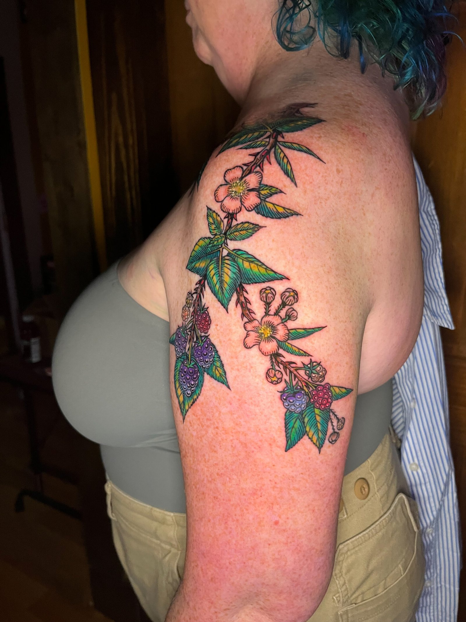A fine line tattoo of a blackberry vine or branch extending from the upper arm to the shoulder of a woman. It has the fruit and flowers of the species surrounded by leaves. This is the Front view. This tattoo was made by Dani Lopez, a tattoo artist at Near Me Tattoo at 6721 Bells Ferry Rd. Ste D110 Woodstock, Ga 30189