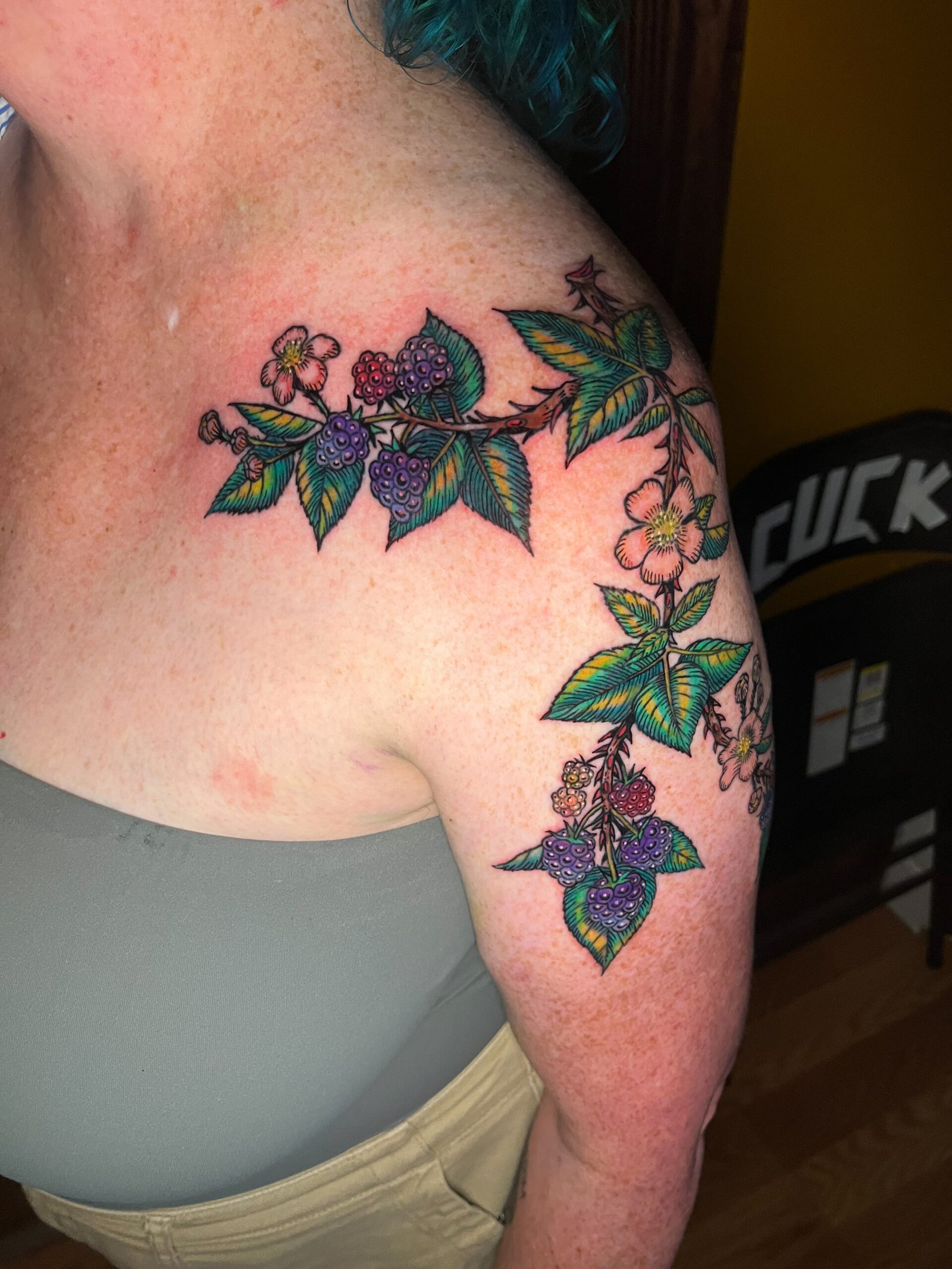 A fine line tattoo of a blackberry vine or branch extending from the upper arm to the shoulder of a woman. It has the fruit and flowers of the species surrounded by leaves. This is the Front view. This tattoo was made by Dani Lopez, a tattoo artist at Near Me Tattoo at 6721 Bells Ferry Rd. Ste D110 Woodstock, Ga 30189