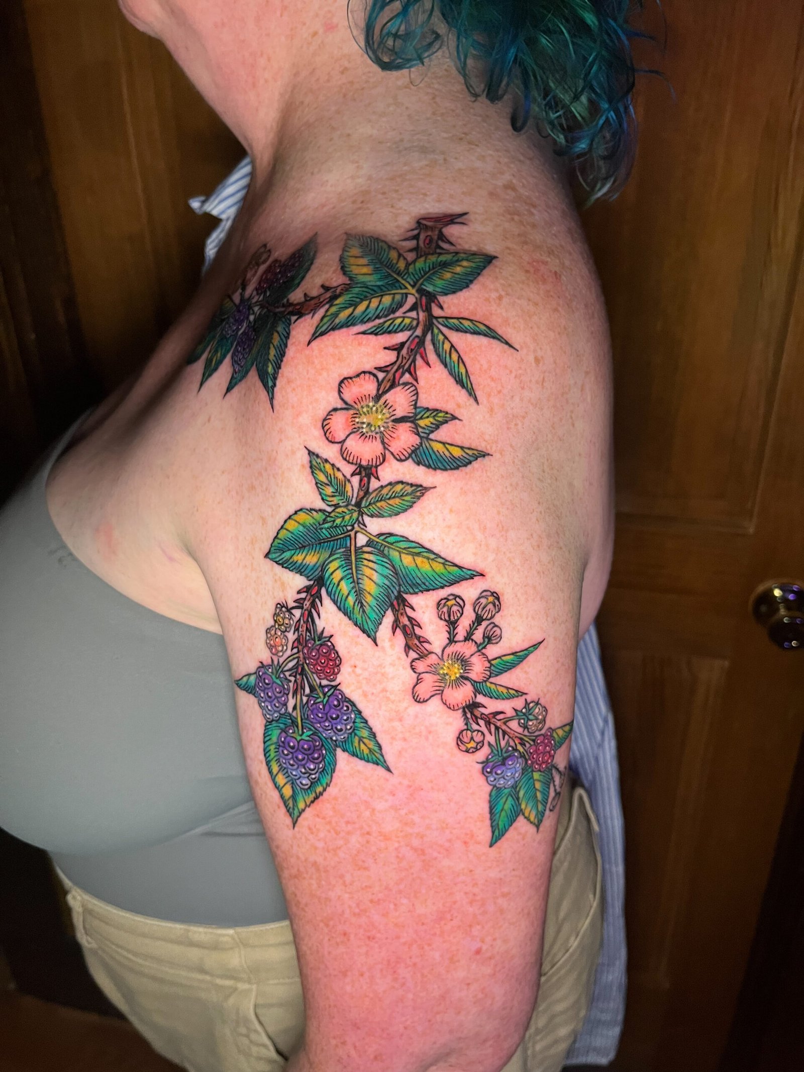 A fine line tattoo of a blackberry vine or branch extending from the upper arm to the shoulder of a woman. It has the fruit and flowers of the species surrounded by leaves. This is the side view. This tattoo was made by Dani Lopez, a tattoo artist at Near Me Tattoo at 6721 Bells Ferry Rd. Ste D110 Woodstock, Ga 30189