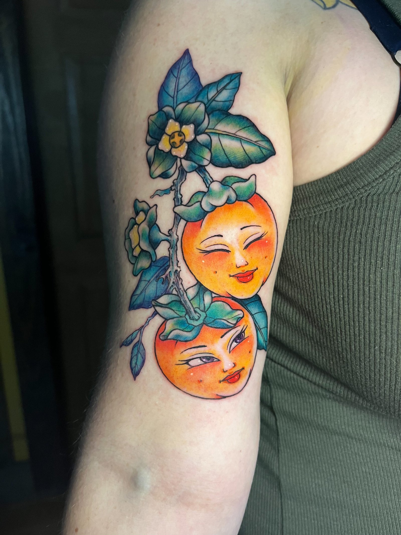 a fine line color Neo traditinonal tattoo of two oranges with smiling feminine faces. One face is smiling with eyes closed. It has deep green leaves and yellow flower buds in it. View 2 This tattoo was made by Dani Lopez, a tattoo artist at Near Me Tattoo at 6721 Bells Ferry Rd. Ste D110 Woodstock, Ga 30189