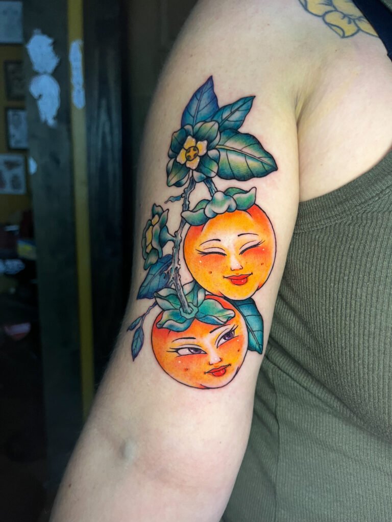 a fine line color Neo traditinonal tattoo of two oranges with smiling feminine faces. One face is smiling with eyes closed. It has deep green leaves and yellow flower buds in it. View 1. This tattoo was made by Dani Lopez, a tattoo artist at Near Me Tattoo at 6721 Bells Ferry Rd. Ste D110 Woodstock, Ga 30189