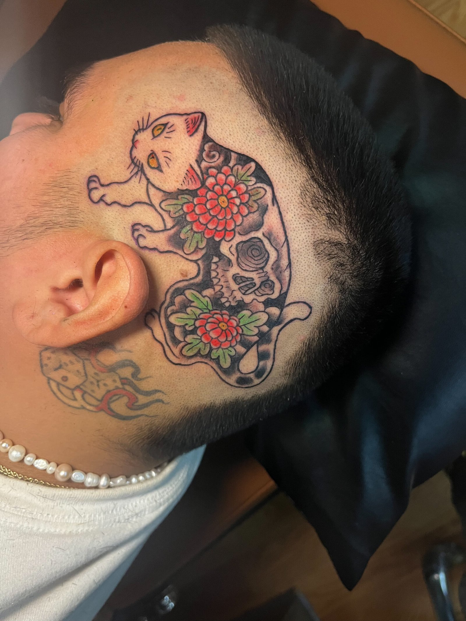 a tattoo done in a Japanese traditional style depicting a cat with a full body tattoo of two small simple chrysanthemums and a skull on its back. The tattoo is located on the temple of a mans head. This tattoo was made by Dani Lopez, a tattoo artist at Near Me Tattoo at 6721 Bells Ferry Rd. Ste D110 Woodstock, Ga 30189