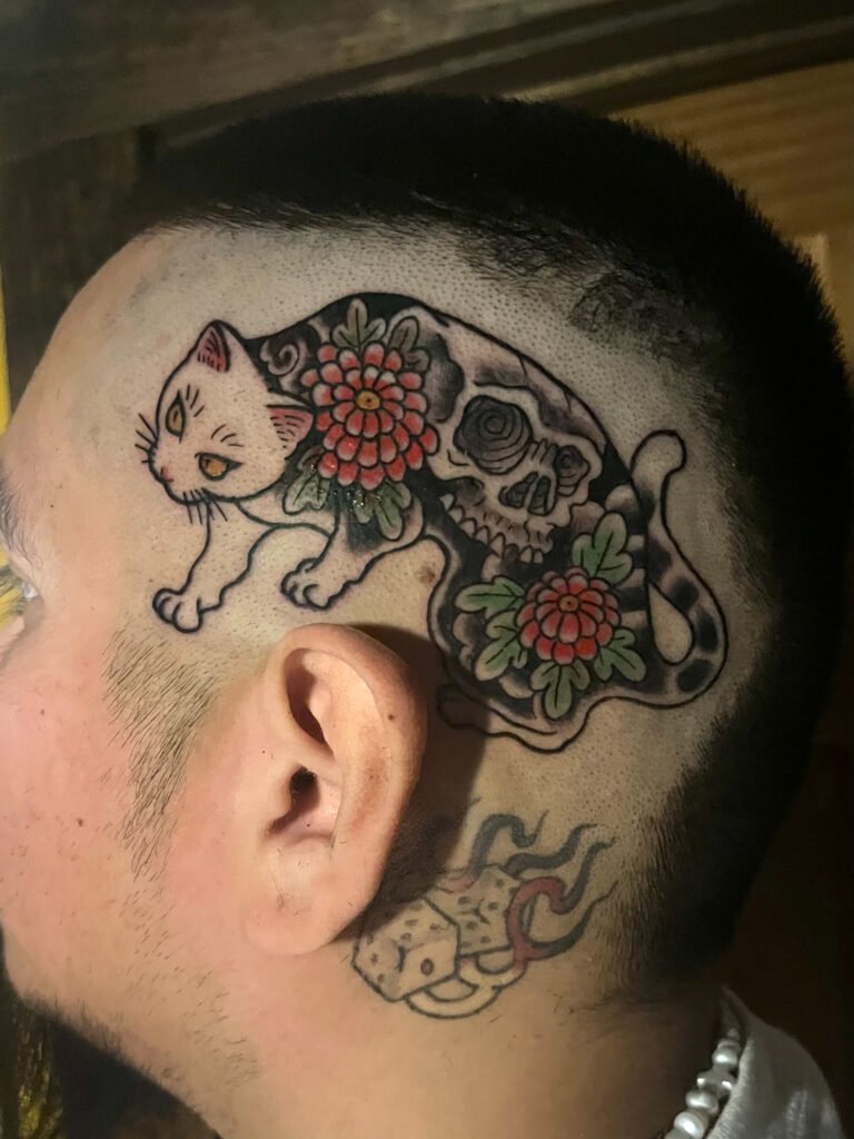 a tattoo done in a Japanese traditional style depicting a cat with a full body tattoo of two small simple chrysanthemums and a skull on its back. The tattoo is located on the temple of a mans head. This tattoo was made by Dani Lopez, a tattoo artist at Near Me Tattoo at 6721 Bells Ferry Rd. Ste D110 Woodstock, Ga 30189 View 2