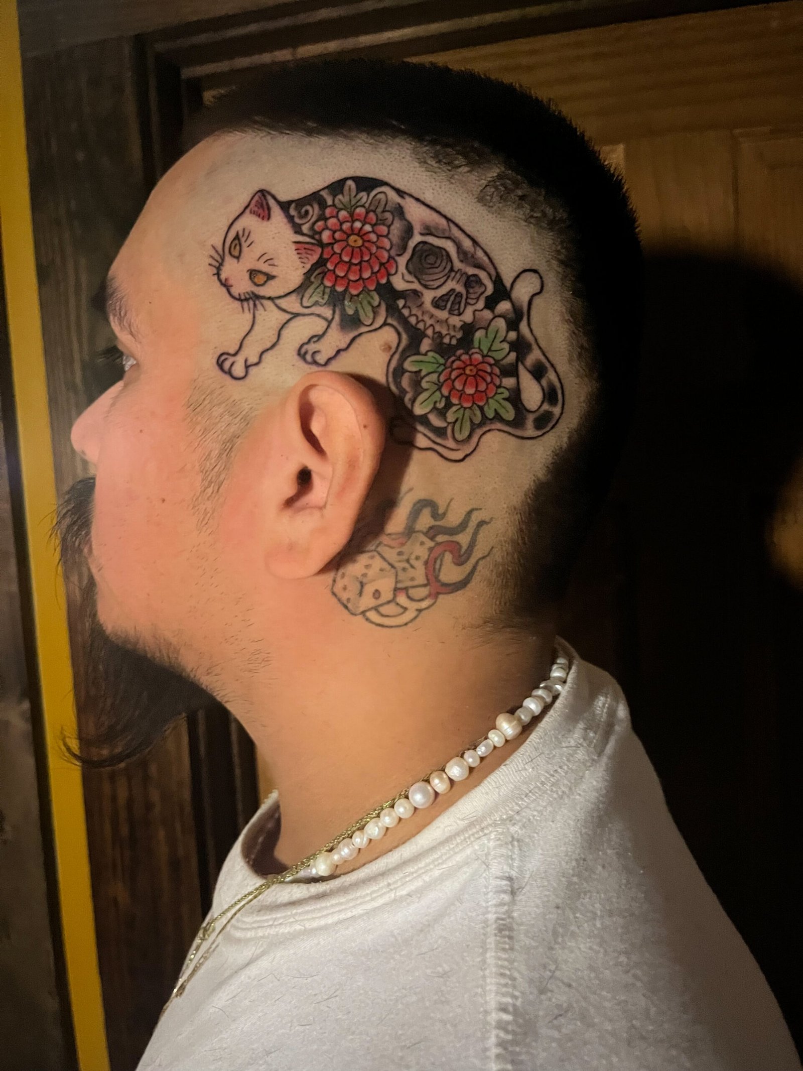 a tattoo done in a Japanese traditional style depicting a cat with a full body tattoo of two small simple chrysanthemums and a skull on its back. The tattoo is located on the temple of a mans head. This tattoo was made by Dani Lopez, a tattoo artist at Near Me Tattoo at 6721 Bells Ferry Rd. Ste D110 Woodstock, Ga 30189 View 3