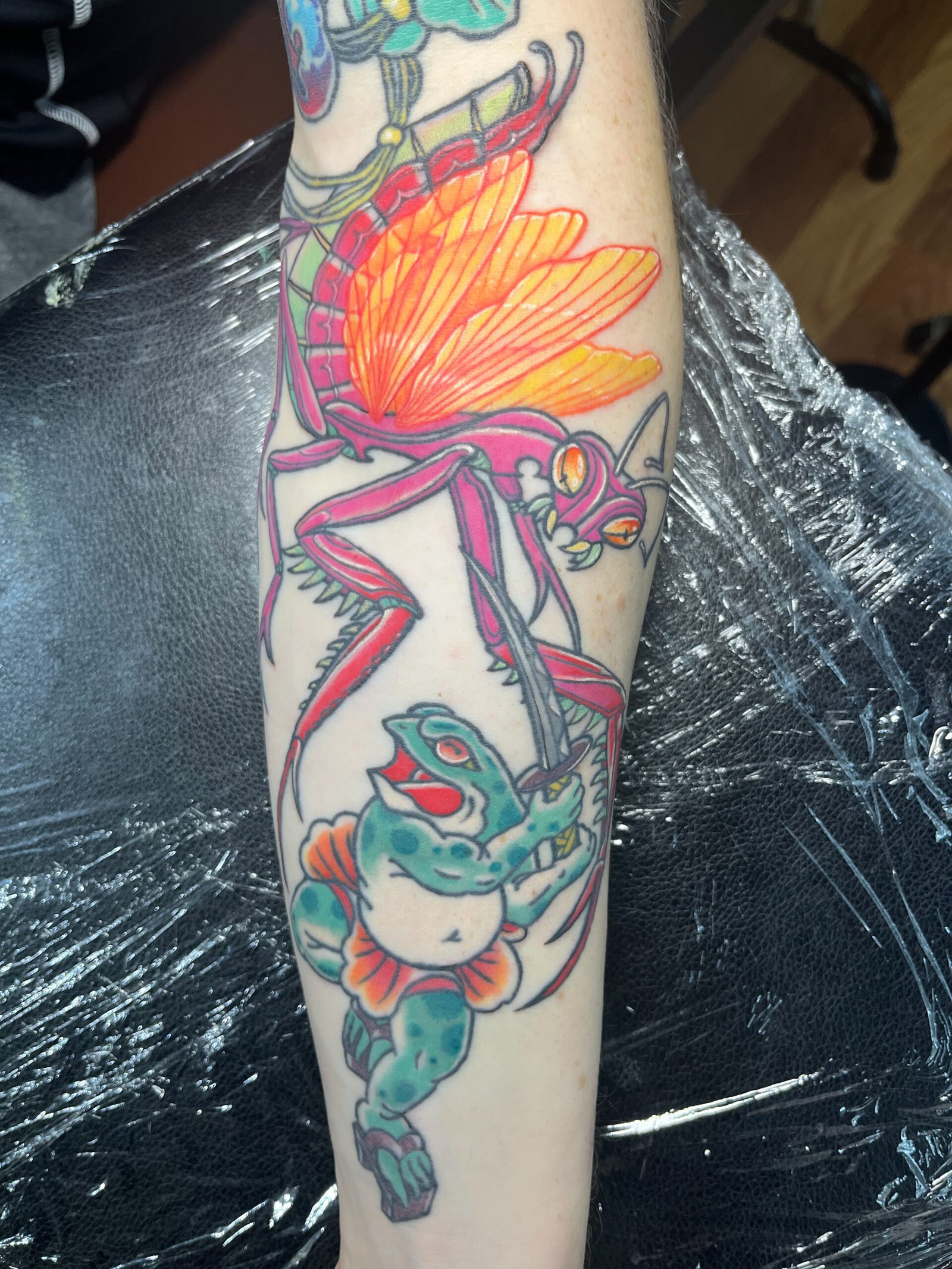 A tattoo done in a mashup of Neo traditional and traditional Japanese styles. It shows a pink praying mantis with glowing yellow and orange wings in battle with a samurai frog wielding a katana sword. It is in a persons forearm. This tattoo was made by Dani Lopez, a tattoo artist at Near Me Tattoo at 6721 Bells Ferry Rd. Ste D110 Woodstock, Ga 30189 View 1