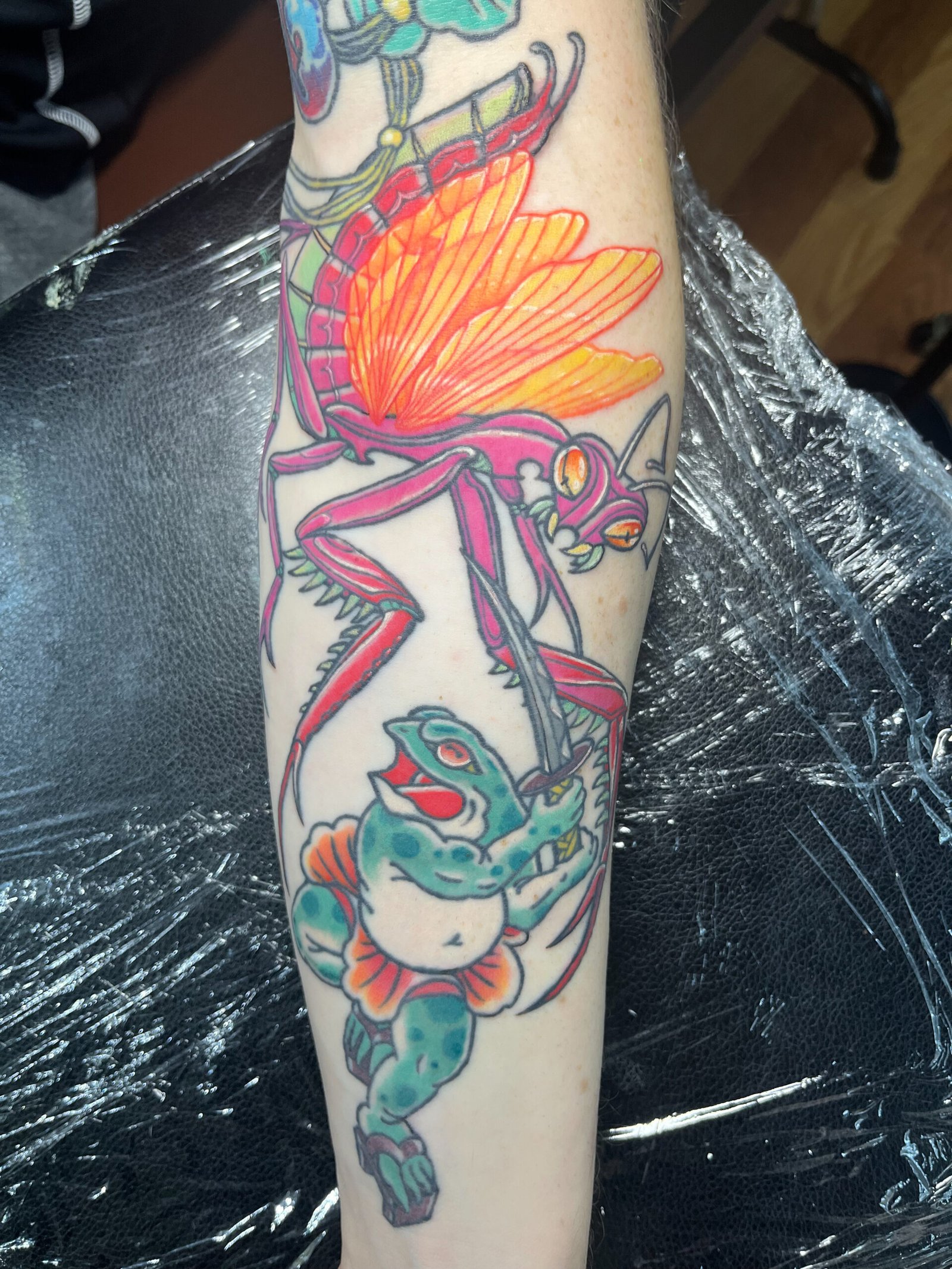 A tattoo done in a mashup of Neo traditional and traditional Japanese styles. It shows a pink praying mantis with glowing yellow and orange wings in battle with a samurai frog wielding a katana sword. It is in a persons forearm. This tattoo was made by Dani Lopez, a tattoo artist at Near Me Tattoo at 6721 Bells Ferry Rd. Ste D110 Woodstock, Ga 30189 View 2
