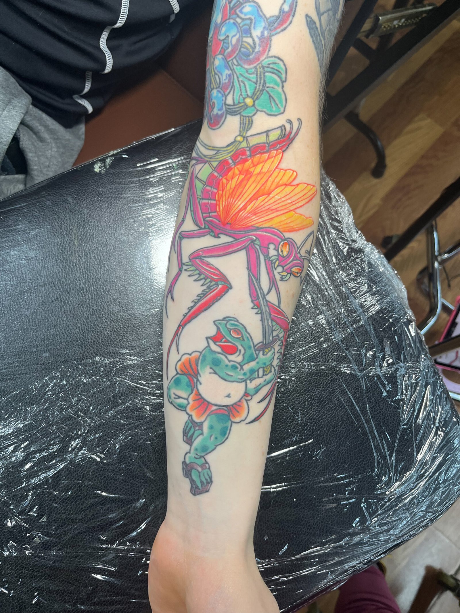 A tattoo done in a mashup of Neo traditional and traditional Japanese styles. It shows a pink praying mantis with glowing yellow and orange wings in battle with a samurai frog wielding a katana sword. It is in a persons forearm. This tattoo was made by Dani Lopez, a tattoo artist at Near Me Tattoo at 6721 Bells Ferry Rd. Ste D110 Woodstock, Ga 30189 view 3