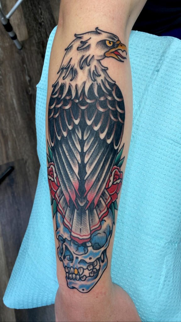 A Neo traditional style eagle sitting on top of a skull with two roses. The eagle feather texture is done with clean whip shading and you can see the eagles wings as they are tucked closely to the body. The eagles head is turned in profile view with fierce eyes. The skull is blue with a gold tooth. This tattoo was done by Jeremy Moffitt an artist at Near Me Tattoo located at 6721 Bells Ferry Rd Ste D110 Woodstock, Ga. 30189