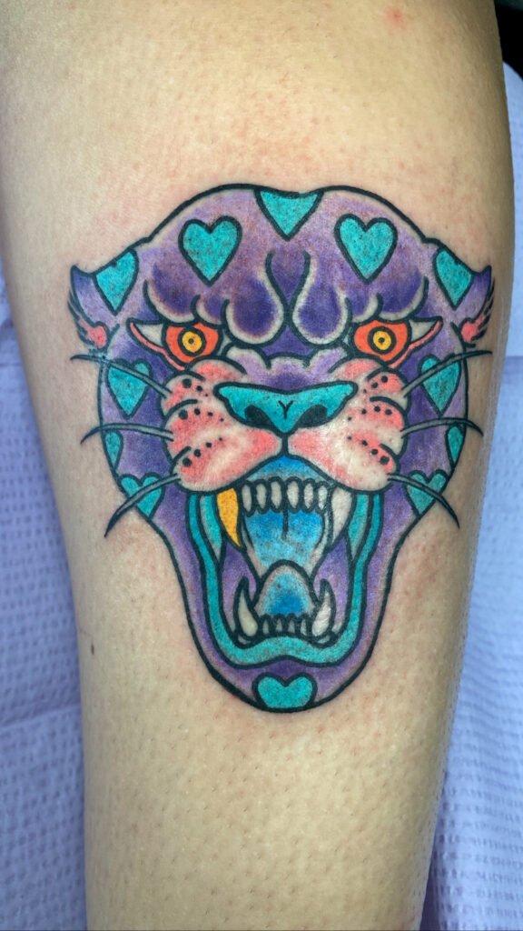 A Neo traditional take on an American traditional snarling panther head tattoo. It is comprised of cool colored inks and the panthers spots are blue hearts. one of the panthers fang teeth is a gold one. It is one of two matching tattoos of this design. This tattoo was done by Jeremy Moffitt an artist at Near Me Tattoo located at 6721 Bells Ferry Rd Ste D110 Woodstock, Ga. 30189
