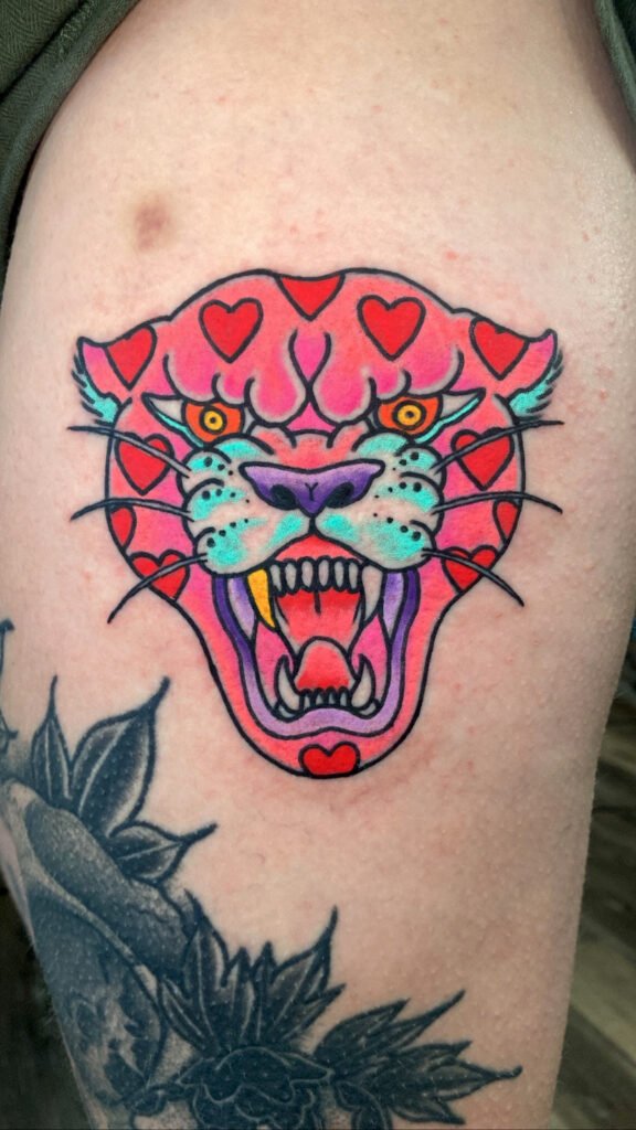 A Neo traditional take on an American traditional snarling panther head tattoo. It is comprised of pink colored inks and the panthers spots are red hearts. one of the panthers fang teeth is a gold one. It is one of two matching tattoos of this design. This tattoo was done by Jeremy Moffitt an artist at Near Me Tattoo located at 6721 Bells Ferry Rd Ste D110 Woodstock, Ga. 30189
