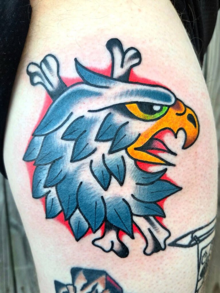 a Neo traditional eagle tattoo. It has opaque grey for the shading in the white feathered and a bright yellow beak with a golden eye with the green glint that eagles have. It has crossbones behind it and background shading of a bright red.