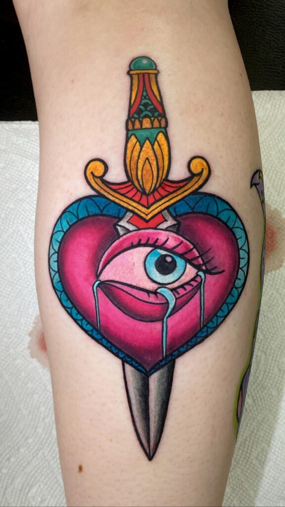 A Neo traditional heart with an eye and an ornate dagger going through it. It is brightly colored and the eye is crying. The design is on the outer shin of the wearer. This tattoo was done by Jeremy Moffitt an artist at Near Me Tattoo located at 6721 Bells Ferry Rd Ste D110 Woodstock, Ga. 30189