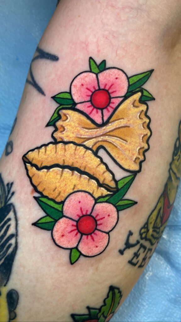 A neo traditional tattoo of bow tie or farfalle and shell or orzo pasta surrounded by simple pink flowers. it shows off a fun culinary food tattoo done with appealing colored ink. This tattoo was done by Jeremy Moffitt an artist at Near Me Tattoo located at 6721 Bells Ferry Rd Ste D110 Woodstock, Ga. 30189