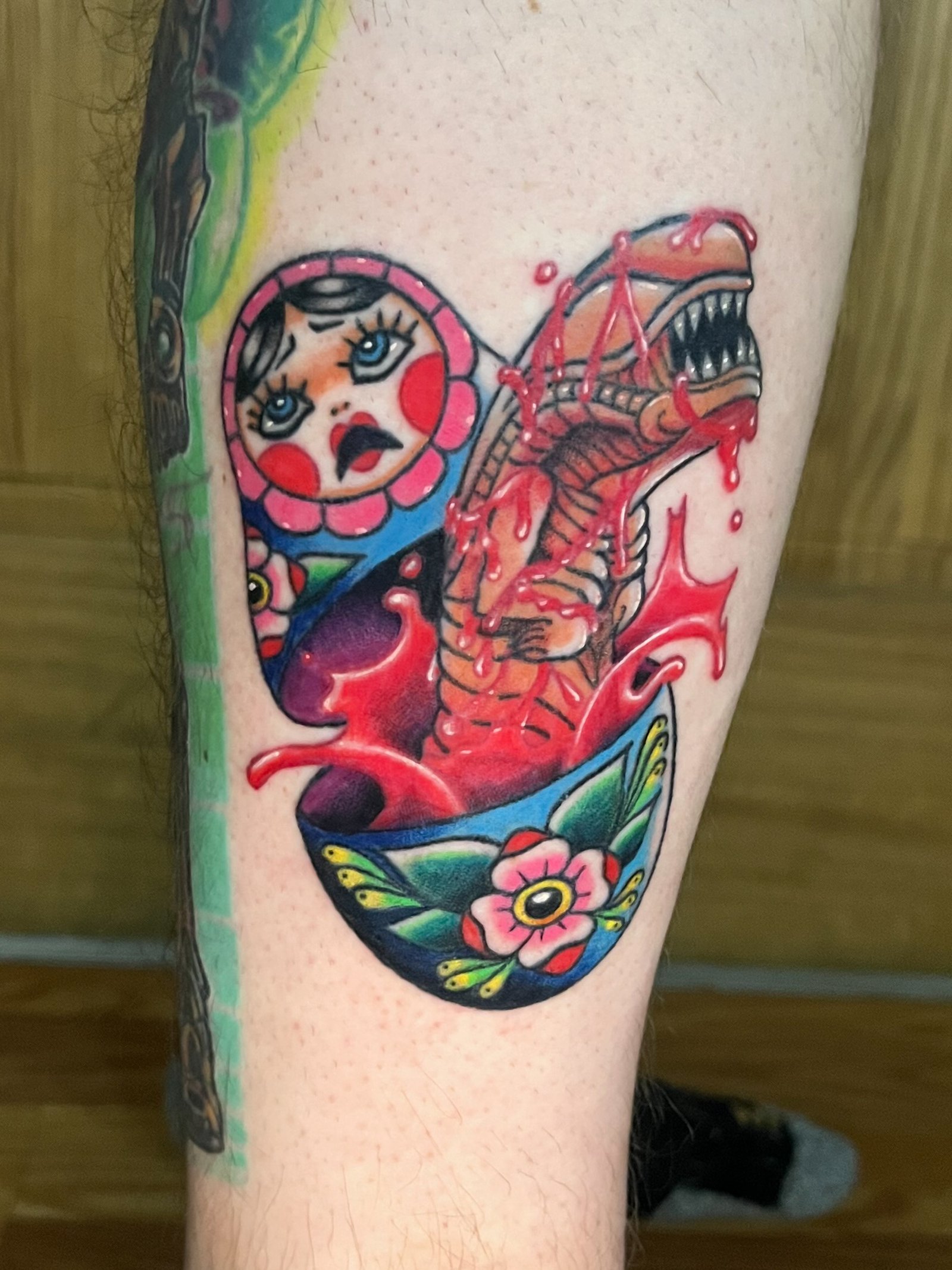 A Neo traditional tattoo mashup tattoo of an alien Xenomorph chestburster popping out of a Russian nesting doll. There is blood colored liquid bursting all around from the inside of the doll and the doll has a shocked look on its face. This tattoo was made by Dani Lopez, a tattoo artist at Near Me Tattoo at 6721 Bells Ferry Rd. Ste D110 Woodstock, Ga 30189