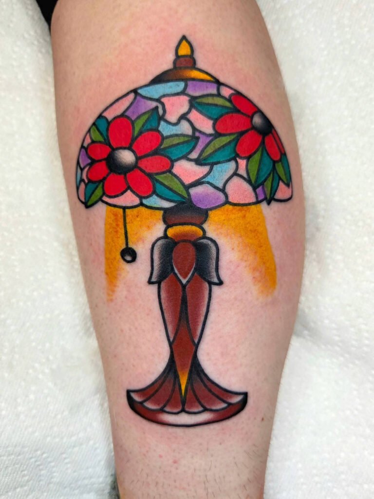 An American traditional tattoo depicting a lit glass domed table lamp. It has a wood colored base and the glass dome depicts stained glass images of red poppy style flowers and blue purple and pink background shapes. This tattoo was made by Noah Beach, co owner of Near Me Tattoo at 6721 Bells Ferry Rd Ste D110 Woodstock, Ga. 30189