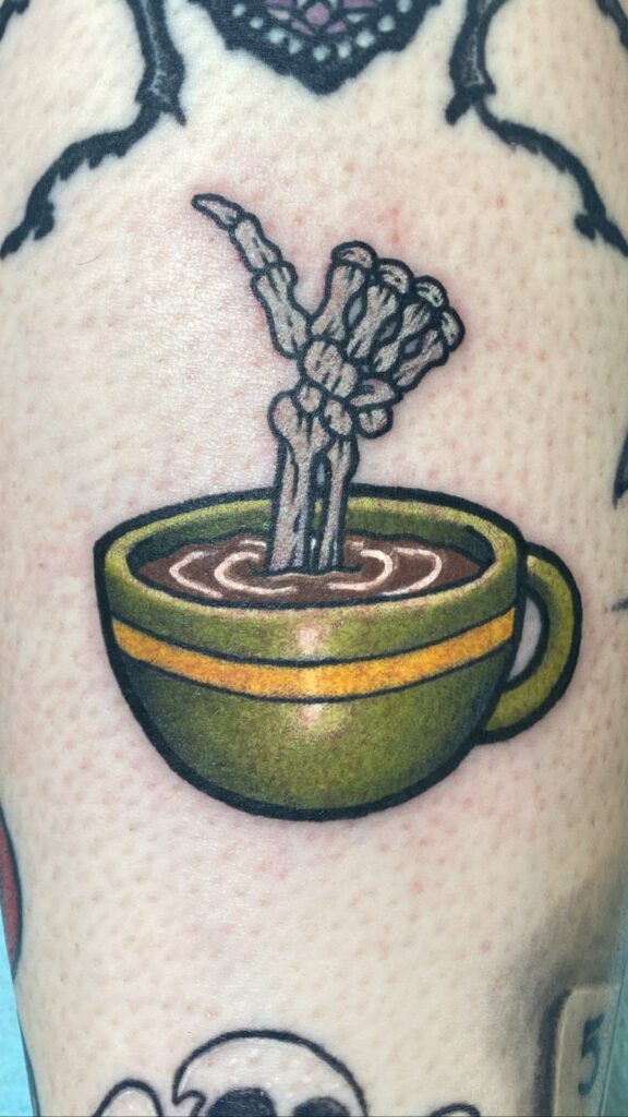 a Neo traditional tattoo of a coffee cup with a skeleton hand coming out of the coffee. The skeletons hand is giving the thumbs up. The mug is an olive green with a golden band. This tattoo was done by Jeremy Moffitt an artist at Near Me Tattoo located at 6721 Bells Ferry Rd Ste D110 Woodstock, Ga. 30189