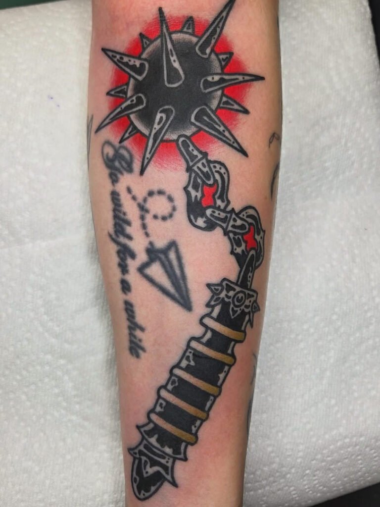 a traditional mace tattoo done in a blackwork shaded style. It has two key colors. Around the handle of the mace are bands of a deep dirty gold color and around the head of the mace ind inside the links of the chain are a vibrant red color. It is on the forearm of the arm and used as a filler piece. This tattoo was made by Noah Beach, co owner of Near Me Tattoo at 6721 Bells Ferry Rd Ste D110 Woodstock, Ga. 30189