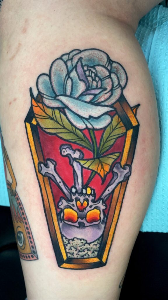 A Neo traditional coffin with skull and blue rose. The skull is upturned with glowing warm yellow orange eyes and a single gold tooth. Crossbones are behind the skull. This tattoo was done by Jeremy Moffitt an artist at Near Me Tattoo located at 6721 Bells Ferry Rd Ste D110 Woodstock, Ga. 30189
