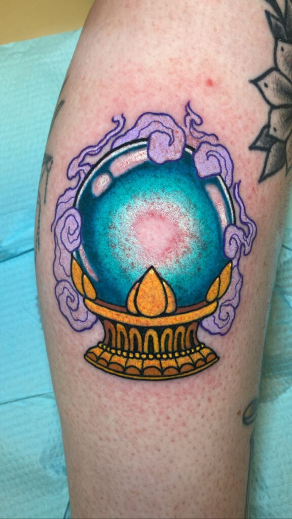 A Neo traditional crystal ball tattoo. The orb of the crystal ball is deep and has a glowing center. The ball sits upon an ornate golden pedestal. There is light purple smoke swirling around the design adding to a mystical aura. This tattoo was done by Jeremy Moffitt an artist at Near Me Tattoo located at 6721 Bells Ferry Rd Ste D110 Woodstock, Ga. 30189