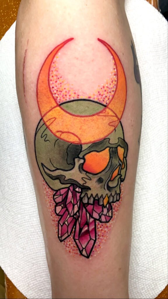 A Neo traditional tattoo of a skull with crystals coming out of its jaw and a crescent moon above that. The skull has glowing orange colored eyes. The background also has a glitter ink effect giving the entire tattoo a colorful and vibrant aura. This tattoo was done by Jeremy Moffitt an artist at Near Me Tattoo located at 6721 Bells Ferry Rd Ste D110 Woodstock, Ga. 30189
