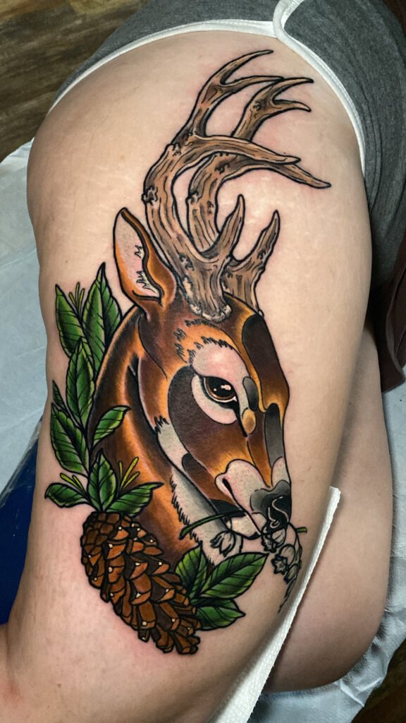 A neo traditional tattoo of a whitetail deer holding a lily of the valley flower sprig in its mouth and framed with a pinecone and leaves at the base of the tattoo. Its placement is on a woman’s thigh. This tattoo was done by Jeremy Moffitt an artist at Near Me Tattoo located at 6721 Bells Ferry Rd Ste D110 Woodstock, Ga. 30189