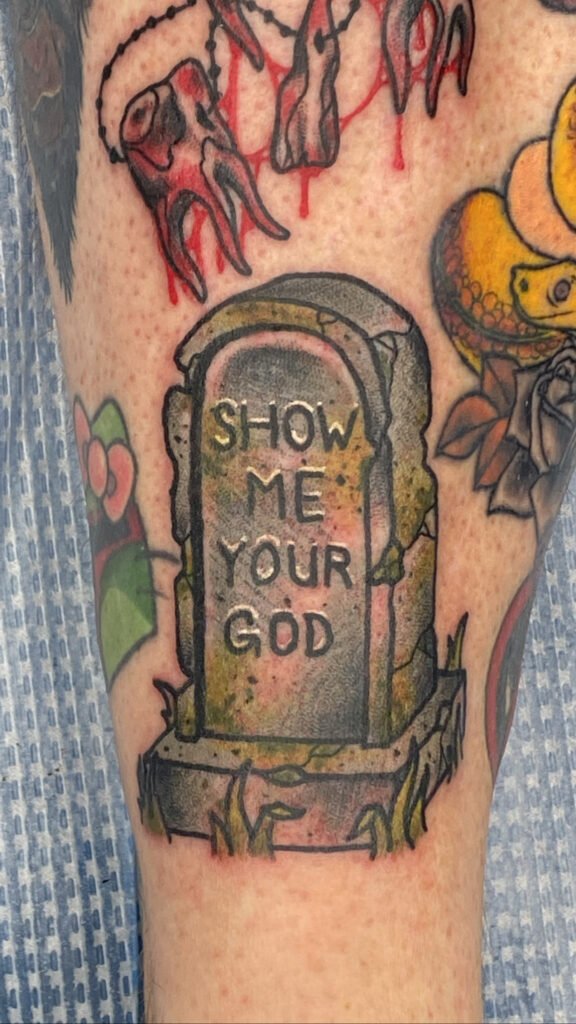 A Neo traditional tattoo of a gravestone. The gravestone is cracked and inscribed on it is “show me your god” . The stone of the headstone is covered in overgrown plants and is mossy looking. This tattoo was done by Jeremy Moffitt an artist at Near Me Tattoo located at 6721 Bells Ferry Rd Ste D110 Woodstock, Ga. 30189