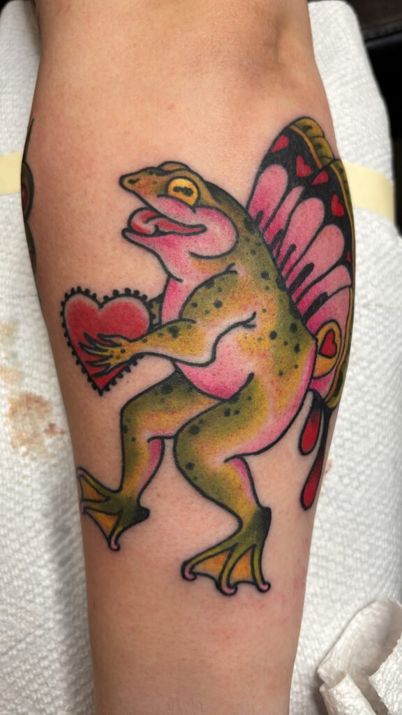 a neo traditional tattoo of a frag standing and holding a heart shape. It has butterfly wings as well that have hearts and are done with predominantly pink ink. This tattoo was done by Jeremy Moffitt an artist at Near Me Tattoo located at 6721 Bells Ferry Rd Ste D110 Woodstock, Ga. 30189