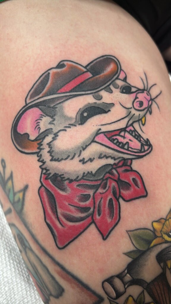 a neo traditional opossum tattoo. The possum is smiling with one gold fang. It is wearing a brown cowboy hat and a handkerchief done in red ink around its neck. This tattoo was done by Jeremy Moffitt an artist at Near Me Tattoo located at 6721 Bells Ferry Rd Ste D110 Woodstock, Ga. 30189