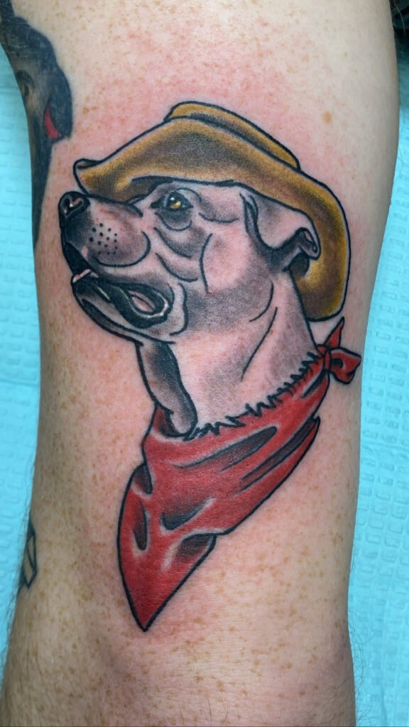 a Neo traditional pet portrait tattoo of a dog wearing a tan cowboy hat and red ink bandanna. The dogs eye is gold and it is hinted with shading that the dog has white fur. This tattoo was done by Jeremy Moffitt an artist at Near Me Tattoo located at 6721 Bells Ferry Rd Ste D110 Woodstock, Ga. 30189