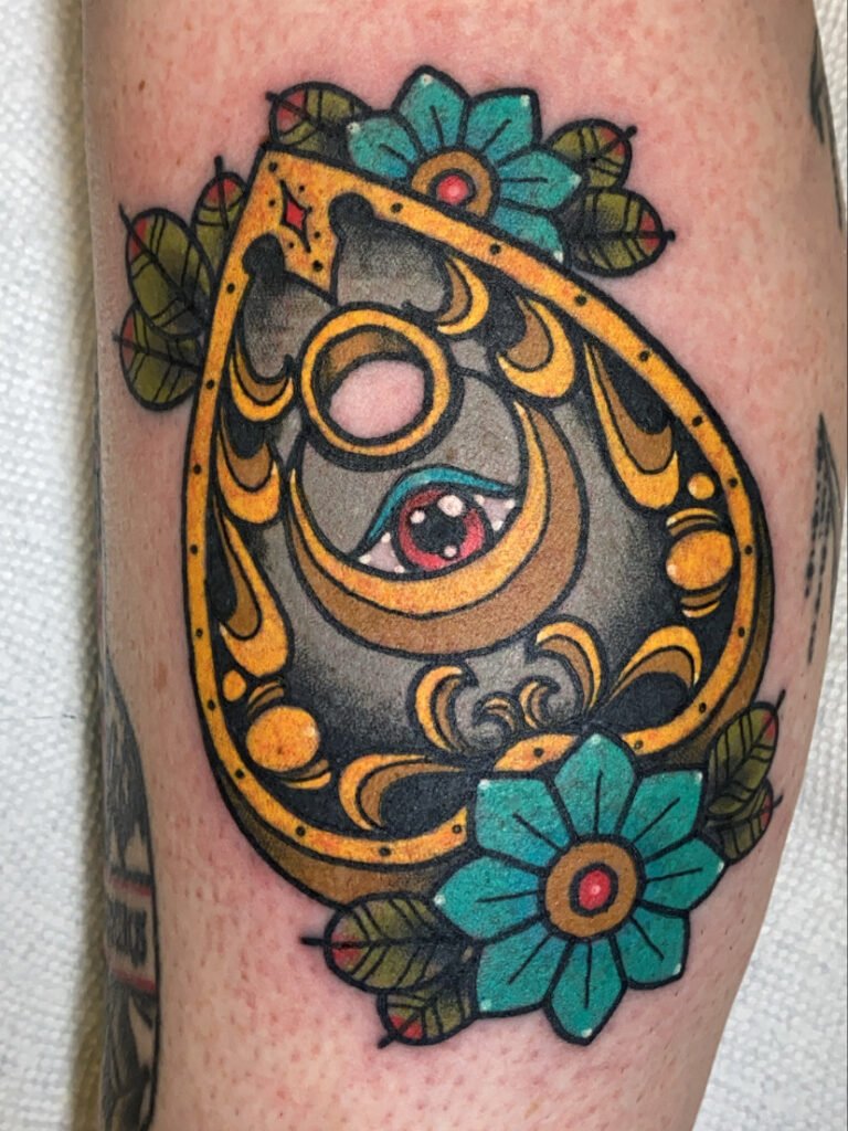 a Neo traditional planchette tattoo. The planchette has an all seeing eye within the ornateness of the design of swooping crescent accents. It has blue ink American traditional flowers. This tattoo was done by Jeremy Moffitt an artist at Near Me Tattoo located at 6721 Bells Ferry Rd Ste D110 Woodstock, Ga. 30189
