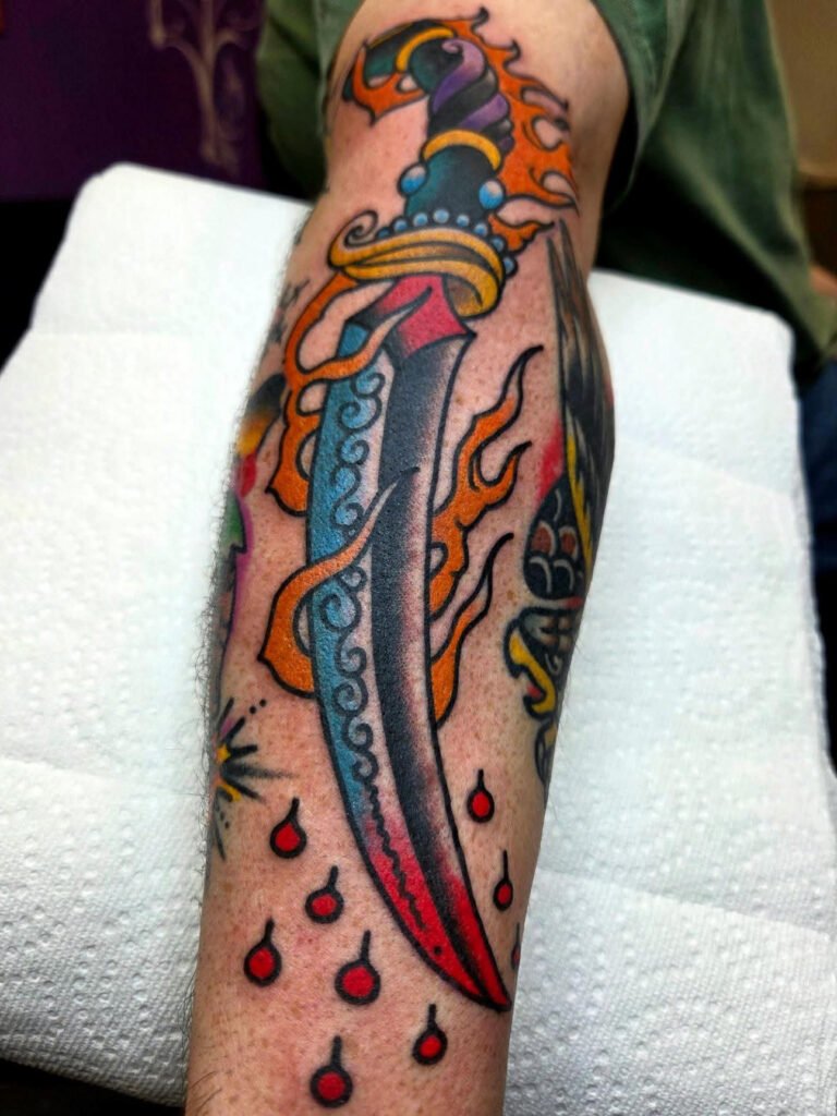 a Neo traditional scimitar sword tattoo. There are orange flames wrapping all the way up the blade and handle. There are blood drops coming of the tip of the sword. This tattoo was made by Noah Beach, co owner of Near Me Tattoo at 6721 Bells Ferry Rd Ste D110 Woodstock, Ga. 30189