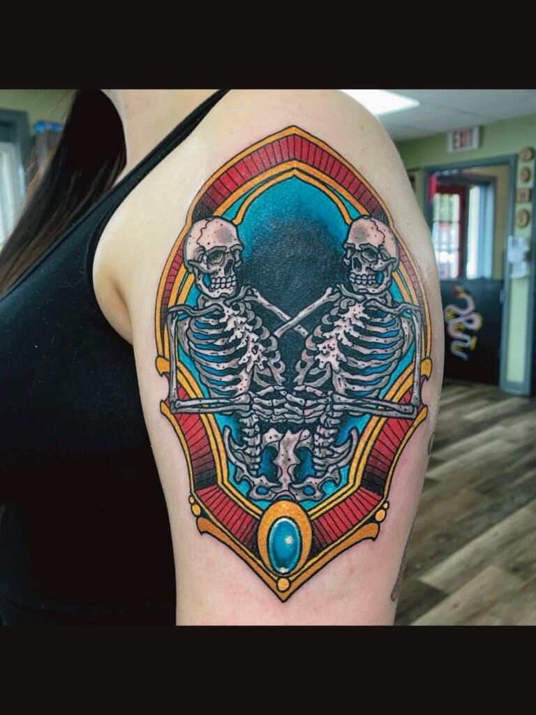 A Neo traditional Siamese skeleton tattoo with an ornate red and gold frame. The skeleton twins are joined at the pelvis and are interlacing their fingers together. This tattoo was done by Jeremy Moffitt an artist at Near Me Tattoo located at 6721 Bells Ferry Rd Ste D110 Woodstock, Ga. 30189