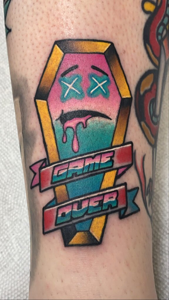A Neo Traditional tattoo of a vaporwave so rad coffin with the text “game over” written in a banner wrapping around the coffin. Inside of the casket is a smiley face with exes for eyes. The colors are magenta pink and teal blues. This tattoo was done by Jeremy Moffitt an artist at Near Me Tattoo located at 6721 Bells Ferry Rd Ste D110 Woodstock, Ga. 30189
