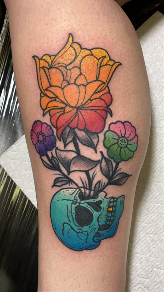 a Neo Traditional tattoo of a skull upturned at the base of the design and three flowers growing from the eye socket. The three flowers are stylized with color faded ink flowing from color to color. This tattoo was done by Jeremy Moffitt an artist at Near Me Tattoo located at 6721 Bells Ferry Rd Ste D110 Woodstock, Ga. 30189