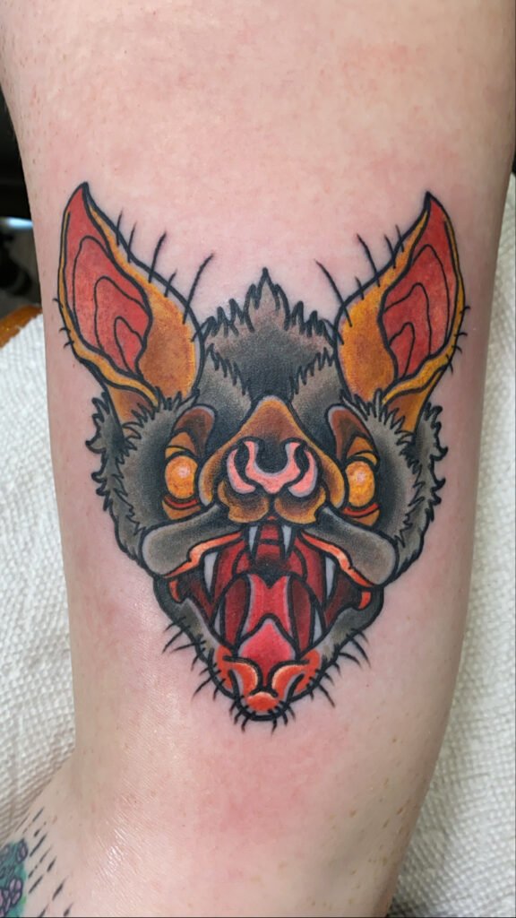 A Neo traditional vampire bat tattoo. It depicts the head of a bat that is looking directly at the viewer. The design has the bat snarling with marbled yellow eyes and long fangs. This tattoo was done by Jeremy Moffitt an artist at Near Me Tattoo located at 6721 Bells Ferry Rd Ste D110 Woodstock, Ga. 30189
