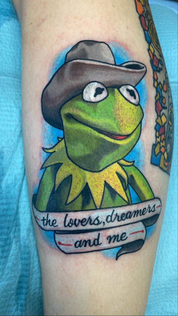 A portrait tattoo of the muppet Kermit the Frog. He is wearing a cowboy hat and on a banner under him it says “the lovers, dreamers, and me” . The design is on the shin of the wearer. This tattoo was done by Jeremy Moffitt an artist at Near Me Tattoo located at 6721 Bells Ferry Rd Ste D110 Woodstock, Ga. 30189