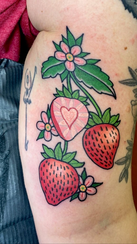 A traditional style tattoo of strawberries on a vine on the upper inner arm tattoo of a woman. One of the three strawberries is cut in half revealing a heart shaped core. On the vine are the pink and white flowers of the strawberry species. This tattoo was done by Jeremy Moffitt an artist at Near Me Tattoo located at 6721 Bells Ferry Rd Ste D110 Woodstock, Ga. 30189