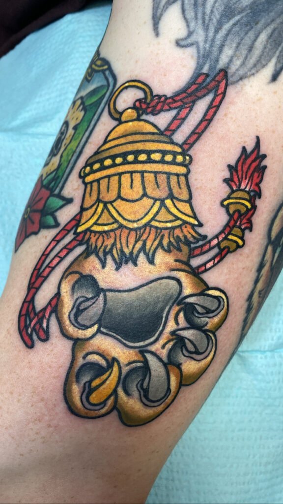 A Neo traditional style Tibetan tiger paw tattoo. It shows the inside pads of the paw with he claws extended out. One of the claws is gold in color. The paw is attached to a golden and ornate cap with red rope going through the loop at the top of the cap. This tattoo was done by Jeremy Moffitt an artist at Near Me Tattoo located at 6721 Bells Ferry Rd Ste D110 Woodstock, Ga. 30189