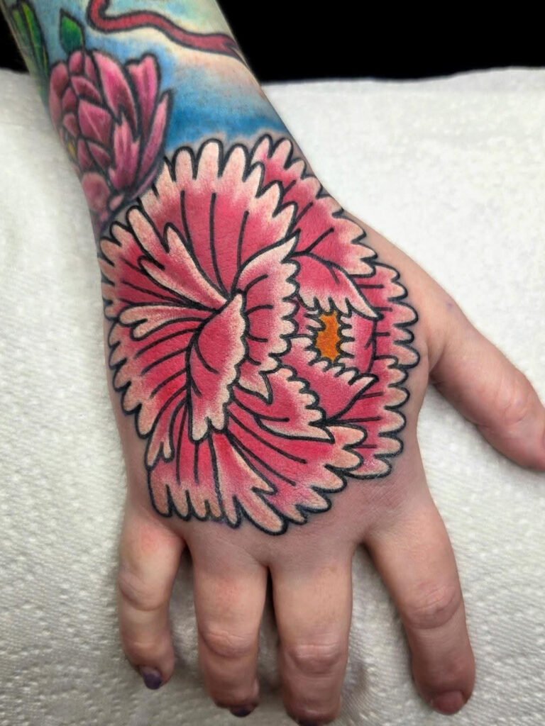 a traditional Japanese Peony on the top of a hand. The petals of the peony are pink with fading into white tips. And the center of the peony is a golden yellow color. This tattoo was made by Noah Beach, co owner of Near Me Tattoo at 6721 Bells Ferry Rd Ste D110 Woodstock, Ga. 30189