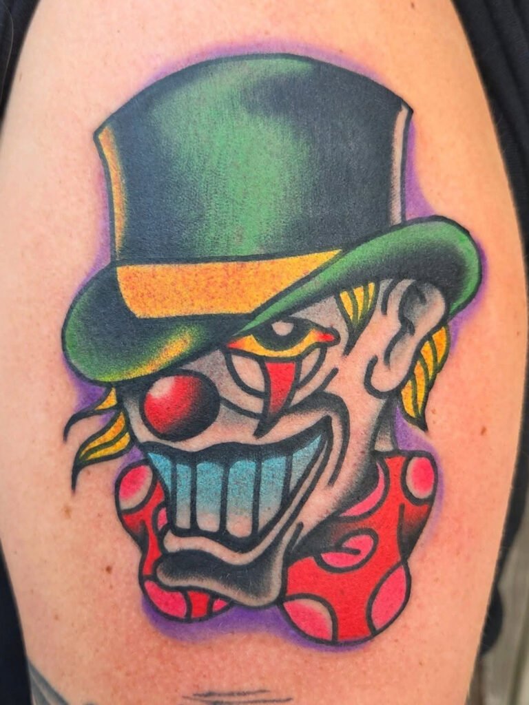 A neo traditional take on a clown ringmaster tattoo. The clown has a large menacing smile with a large green tophat with a yellow brim, the same color as the clowns eyes and hair. The clown has a red nose and bow tie. This tattoo was made by Noah Beach, co owner of Near Me Tattoo at 6721 Bells Ferry Rd Ste D110 Woodstock, Ga. 30189