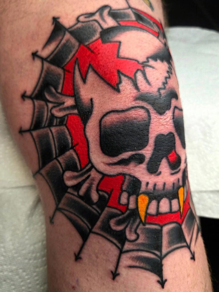 A Neo traditional tattoo on the knee. It depicts a vampire skull with crossbones around it. The vampire skull has golden ranks and it is surrounded in a circular pattern with blackwork spiderwebs.inside of the spiderweb frame and behind the skull is a vibrant red ink color. This tattoo was made by Noah Beach, co owner of Near Me Tattoo at 6721 Bells Ferry Rd Ste D110 Woodstock, Ga. 30189