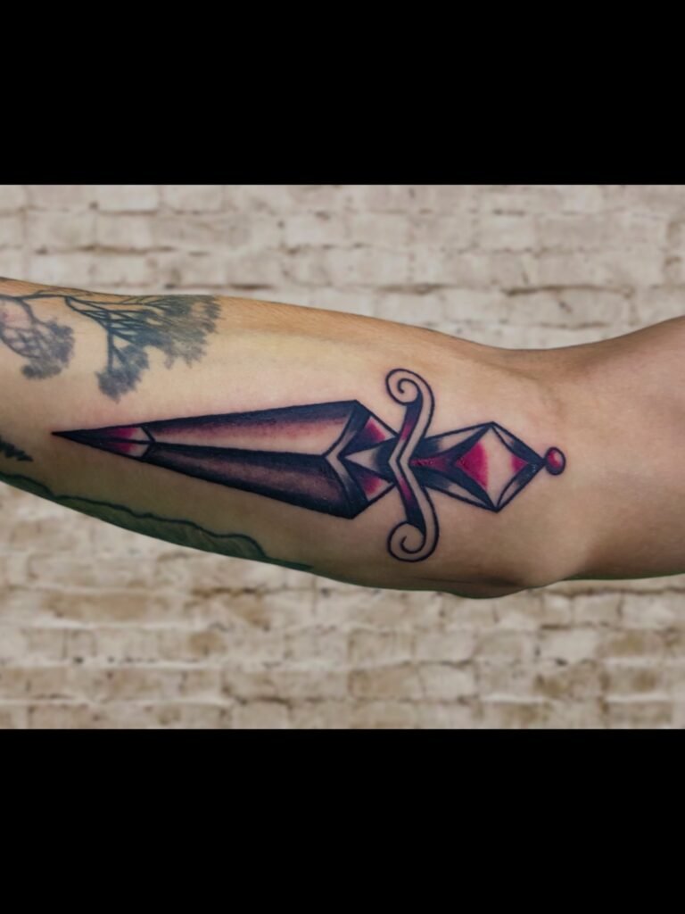 A tattoo of an American Traditional dagger done in black and pomegranate ink. The tip of the blade is pointed toward the hand and finger. It is placed on the inner forearm of wearer. This tattoo was made by Nathan Heinrich, co owner of Near Me Tattoo at 6721 Bells Ferry Rd Ste D110 Woodstock, Ga. 30189