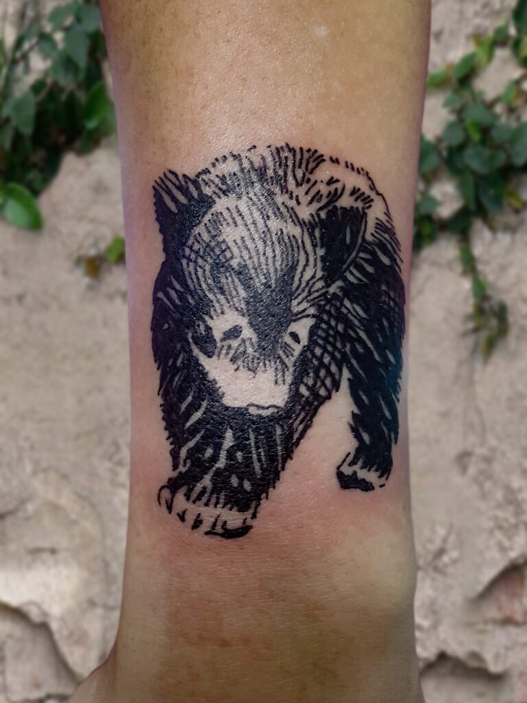 A tattoo of a blackwork illustrative baby black bear cub. It is on the forearm of the wearer. It is a full body 3/4 view of the animal with its paws pointed towards the hand and finger of the wearer. It is a cute simple tattoo that is very feminine. This tattoo was made by Nathan Heinrich, co owner of Near Me Tattoo at 6721 Bells Ferry Rd Ste D110 Woodstock, Ga. 30189