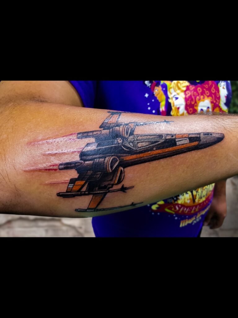 A tattoo of Poe Dameron’s X-wing space fighter ship from the Star Wars franchise. It shows the ship on the wearers forearm at an almost profile angle with the front of the ship pointed toward the hand and finger. The ship is black and grey and orange with bright red jets coming from the engines. This tattoo was made by Nathan Heinrich, co owner of Near Me Tattoo at 6721 Bells Ferry Rd Ste D110 Woodstock, Ga. 30189